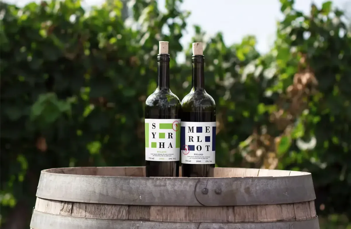 Two corked wine bottles labelled Syrah and Merlot standing on a wooden barrel with a blurred vineyard background.