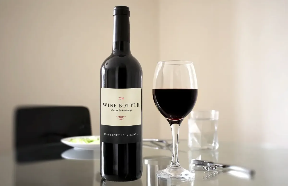 Red wine bottle labelled Wine Bottle Cabernet Sauvignon beside a poured glass of red wine on a dining table, with a corkscrew nearby.