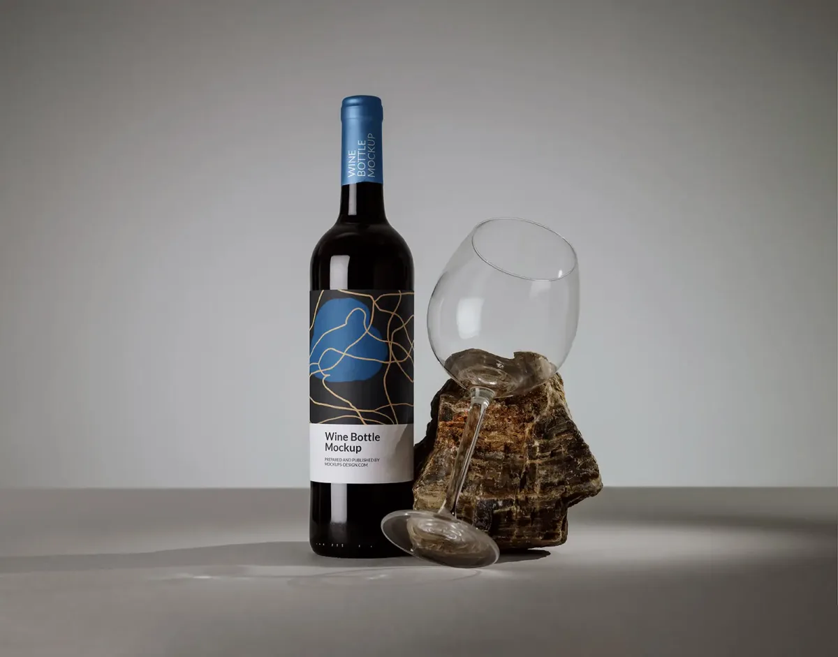 Wine bottle with modern abstract label beside an oversized empty wine glass tilted against a piece of petrified wood, grey studio background.