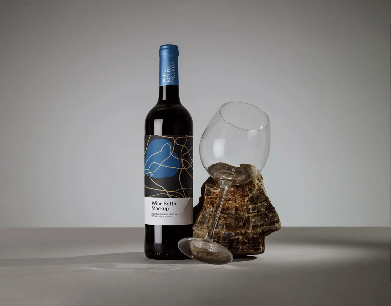 Wine bottle with modern abstract label beside an oversized empty wine glass tilted against a piece of petrified wood, grey studio background.